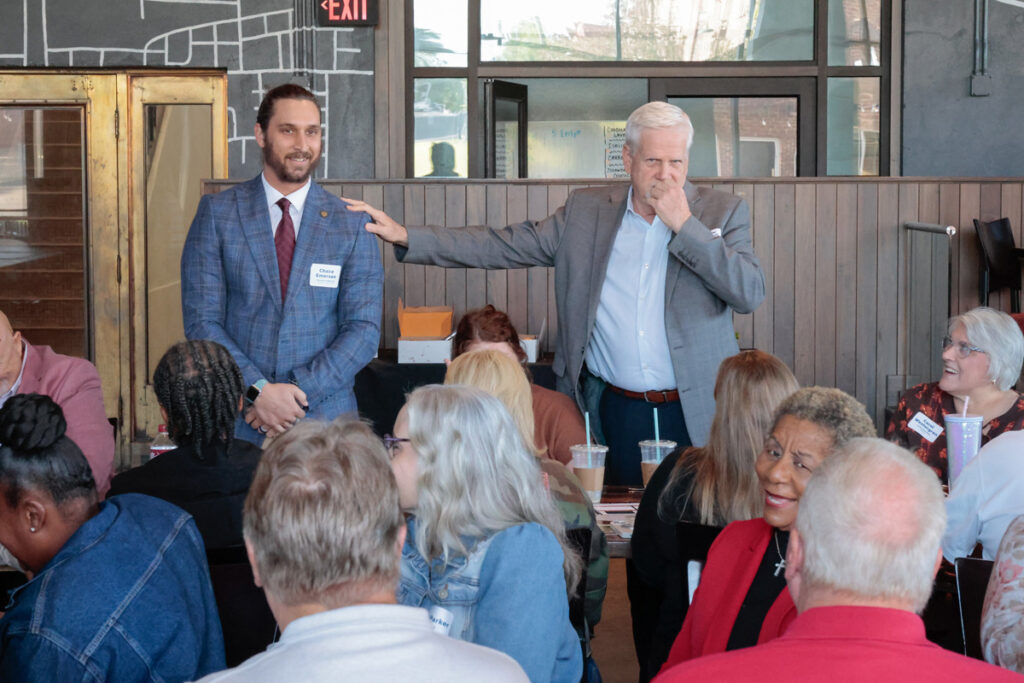 Two speakers stand up in front of a group of meeting attendees.